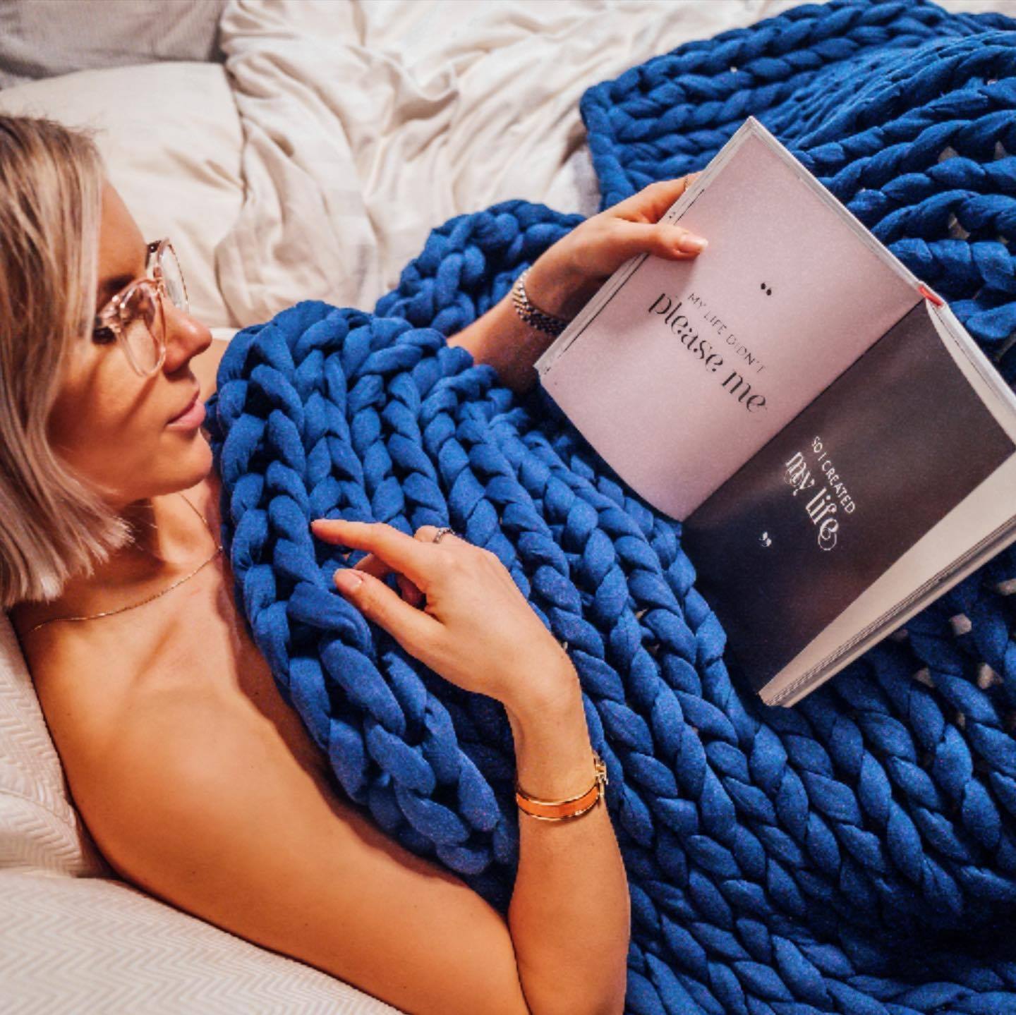 A woman reads a book in bed in a blue knitted weighted blanket